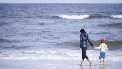 Ein Mutter hält ihre Tochter an der Hand und spaziert mit ihrem am Strand entlang. Im Hintergrund ist das weiter Meer zu sehen.