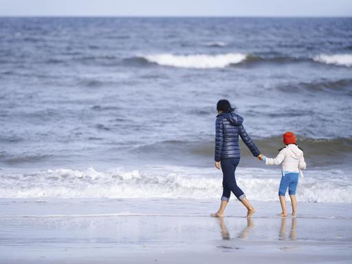 Ein Mutter hält ihre Tochter an der Hand und spaziert mit ihrem am Strand entlang. Im Hintergrund ist das weiter Meer zu sehen.