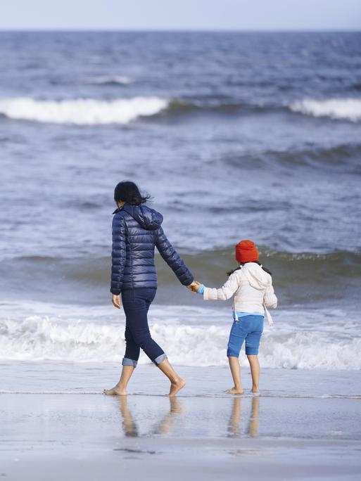 Ein Mutter hält ihre Tochter an der Hand und spaziert mit ihrem am Strand entlang. Im Hintergrund ist das weiter Meer zu sehen.