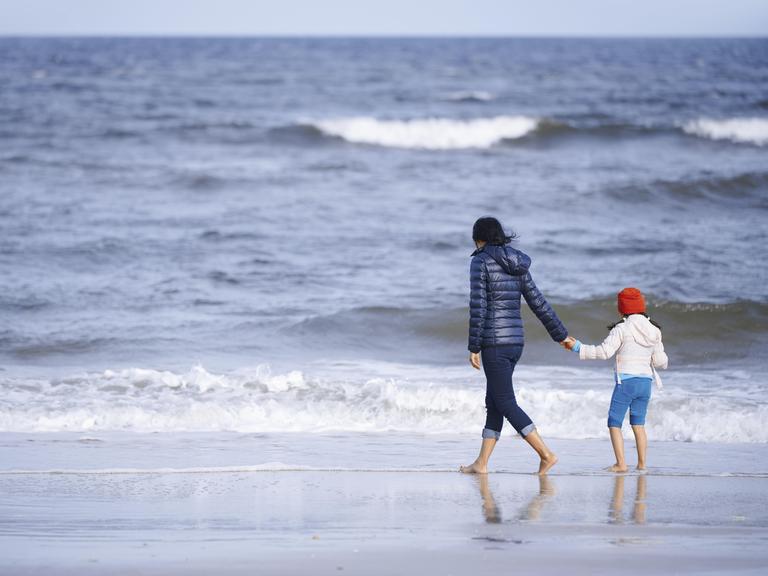 Ein Mutter hält ihre Tochter an der Hand und spaziert mit ihrem am Strand entlang. Im Hintergrund ist das weiter Meer zu sehen.