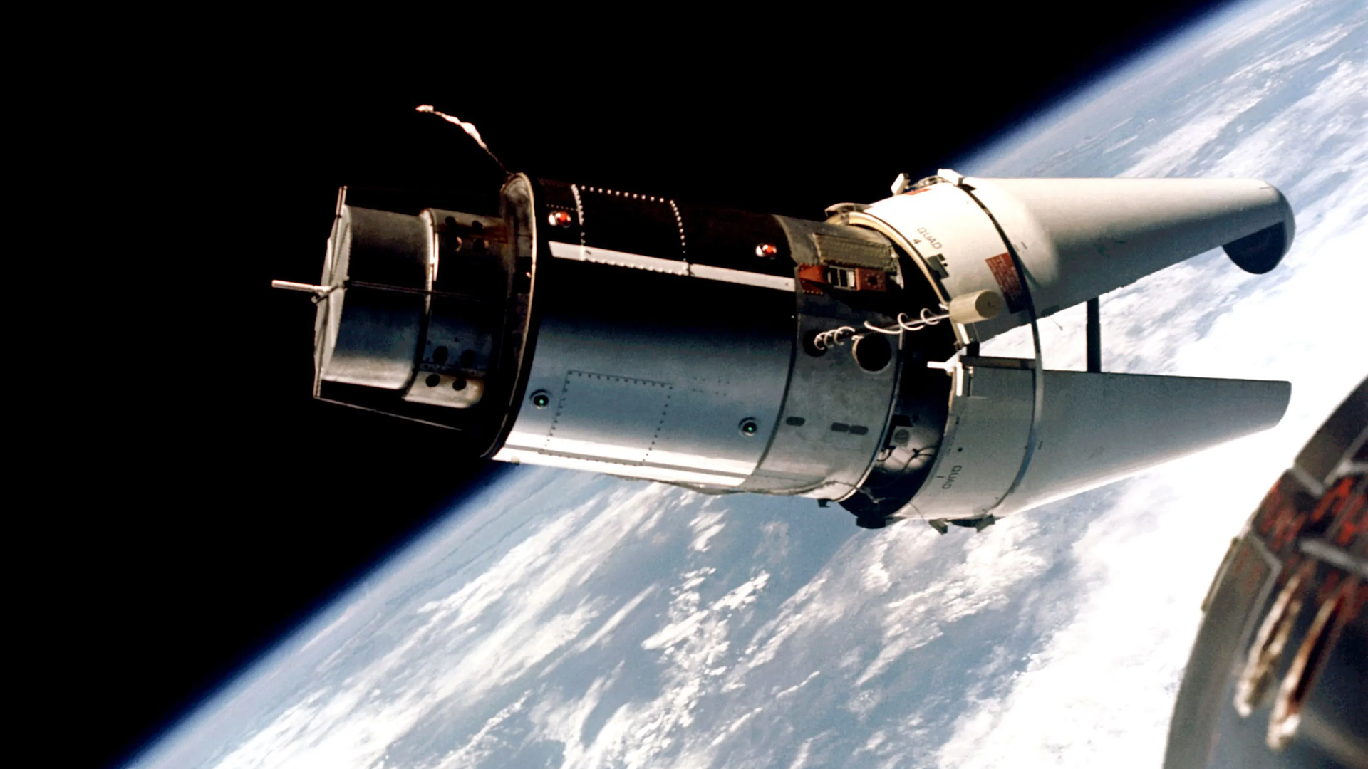 Das Raumschiff Gemini 9 befindet sich im Weltraum über der Erde. Der vordere Teil des Raumschiffs ist geöffnet und sieht dabei so aus, wie das Maul eines Krokodils.