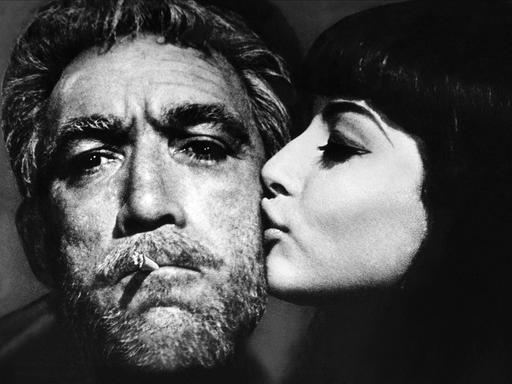 Der Schauspieler Anthony Quinn mit Schauspielerin Eleni Anousaki im Film "Alexis Sorbas". Er hat eine Zigarette im Mund und wird von ihr auf die bärtige Wange geküsst. 