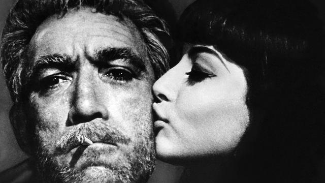 Der Schauspieler Anthony Quinn mit Schauspielerin Eleni Anousaki im Film "Alexis Sorbas". Er hat eine Zigarette im Mund und wird von ihr auf die bärtige Wange geküsst. 