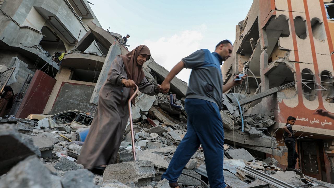 Ein Mann führt eine ältere Frau am Gehstock aus dem Schutt eines zerstörten Gebäudes heraus. Ein Mann führt eine ältere Frau am Gehstock aus dem Schutt eines zerstörten Gebäudes heraus.