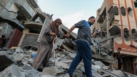 Ein Mann führt eine ältere Frau am Gehstock aus dem Schutt eines zerstörten Gebäudes heraus.