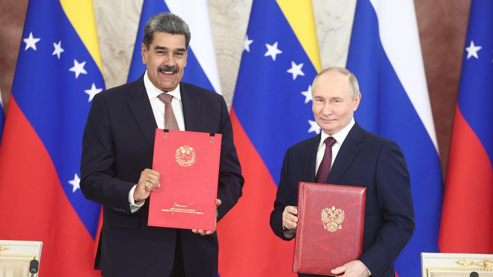 Venezuelas Präsident  Maduro steht lächelnd neben Russlands Präsident Putin vor den Flaggen der beiden Länder. In die Kamera halten die beiden rote Mappen mit goldenen Wappen darauf.