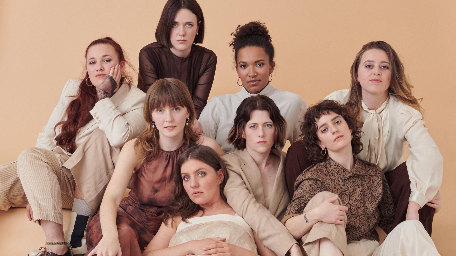 Acht Frauen posieren als Gruppe in beige-brauner Kleidung.