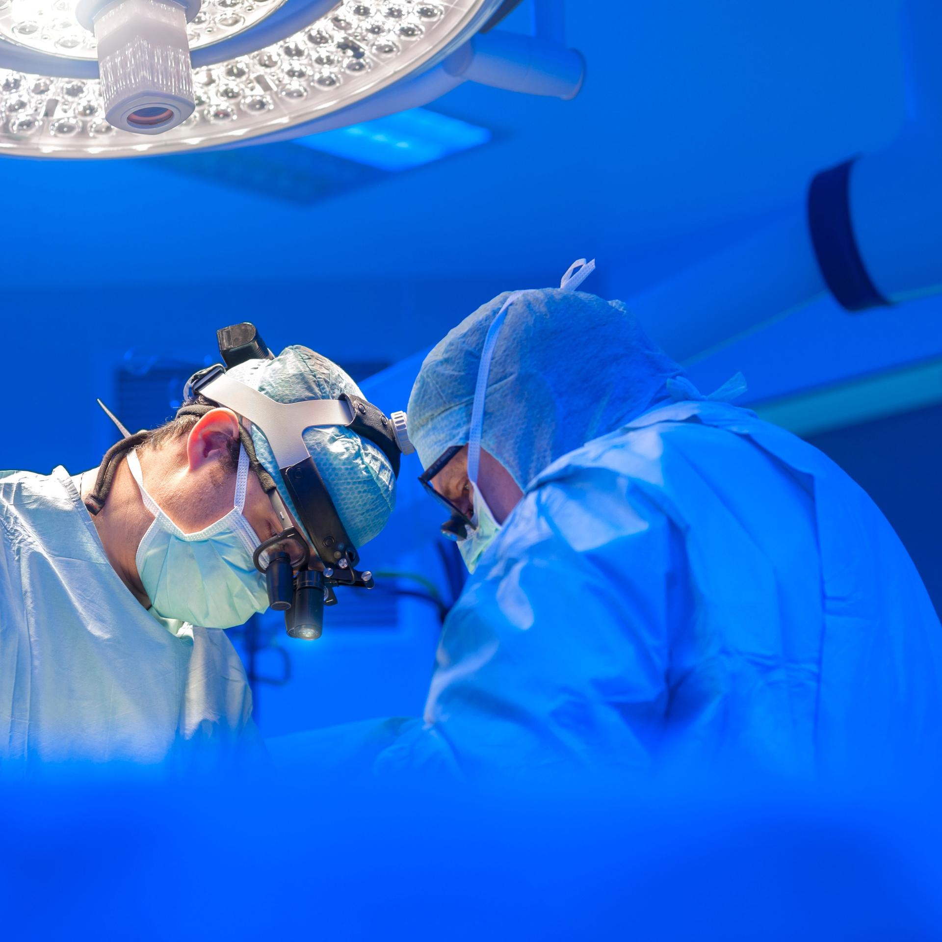 Zwei Ärzte in OP-Kleidung stehen in einem OP-Saal und scheinen zu operieren. Das Licht ist kühl, sie tragen helle Kleidung. 