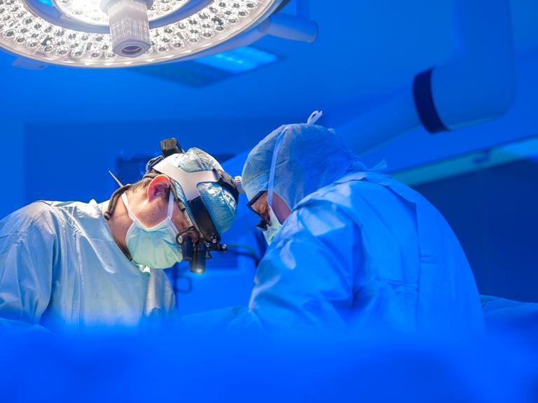 Zwei Ärzte in OP-Kleidung stehen in einem OP-Saal und scheinen zu operieren. Das Licht ist kühl, sie tragen helle Kleidung. 