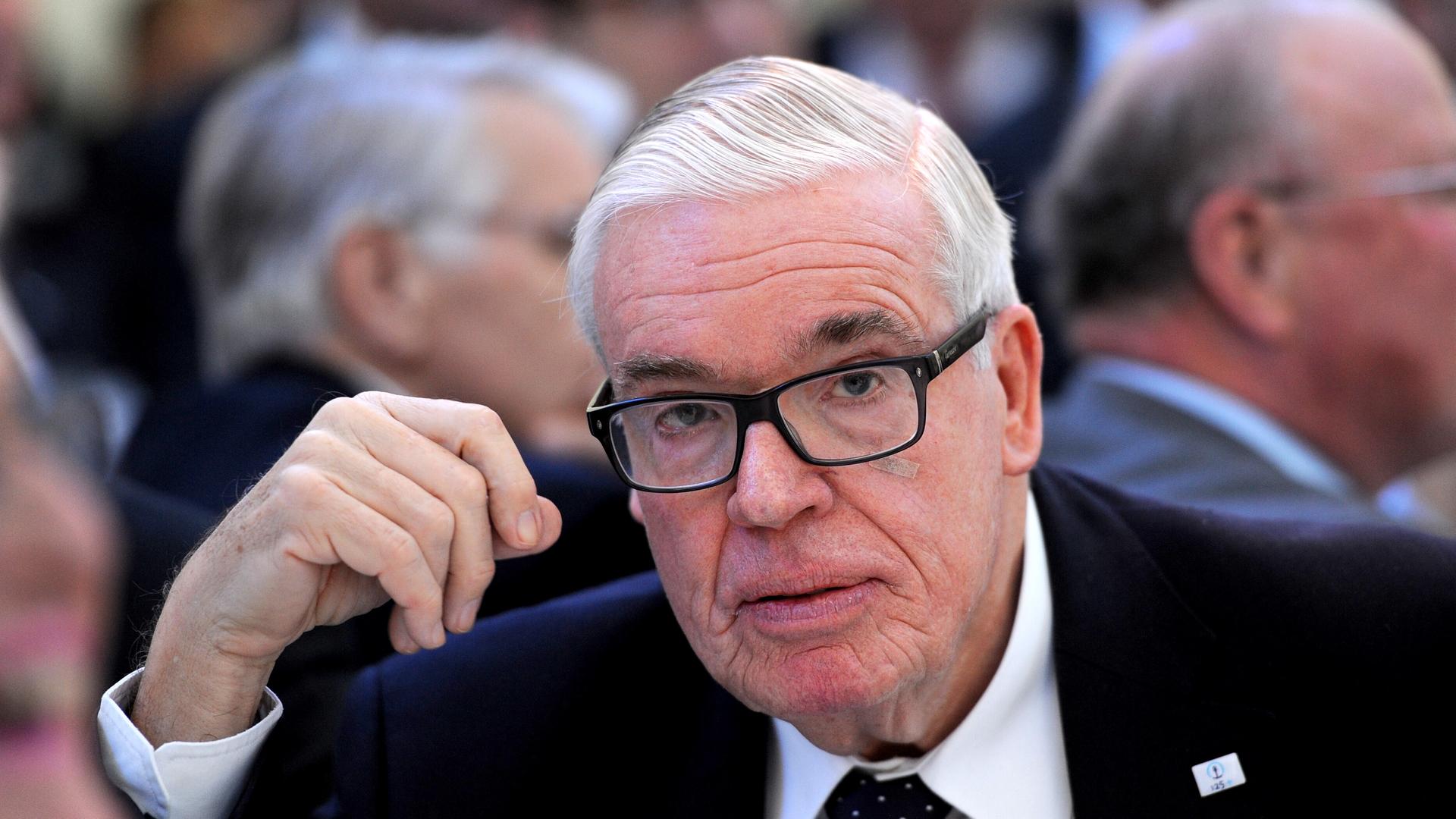 Klaus-Michael Kühne in einem vollbesetzten Saal. Er schaut in die Kamera. Im Hintergrund sitzen Menschen auf Sitzreihen. 