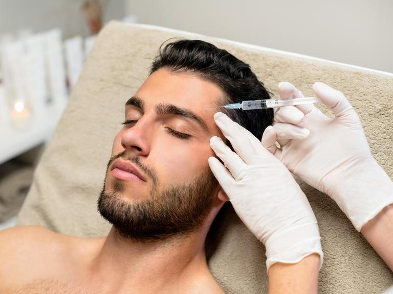 Ein junger Mann liegt auf einer Liege und lässt sich Botox in die Stirn spritzen