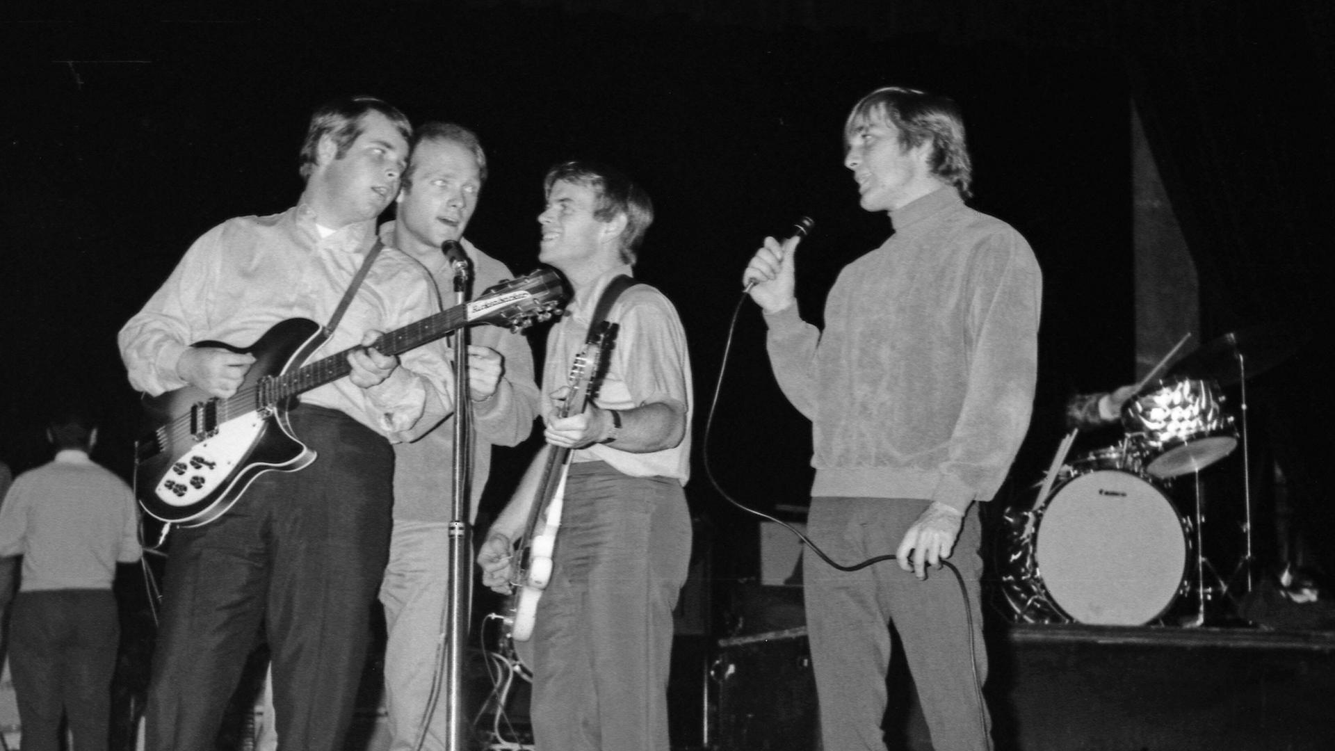 Schwarz-Weiß-Bild von einem Konzert der Band "The Beach Boys" im Jahr 1964 in Paris: Brian Wilson, Mike Love, Al Jardine, Dennis Wilson stehen auf der Bühne dicht beisammen um ein Mikro, Brian Wilson spielt Gitarre.