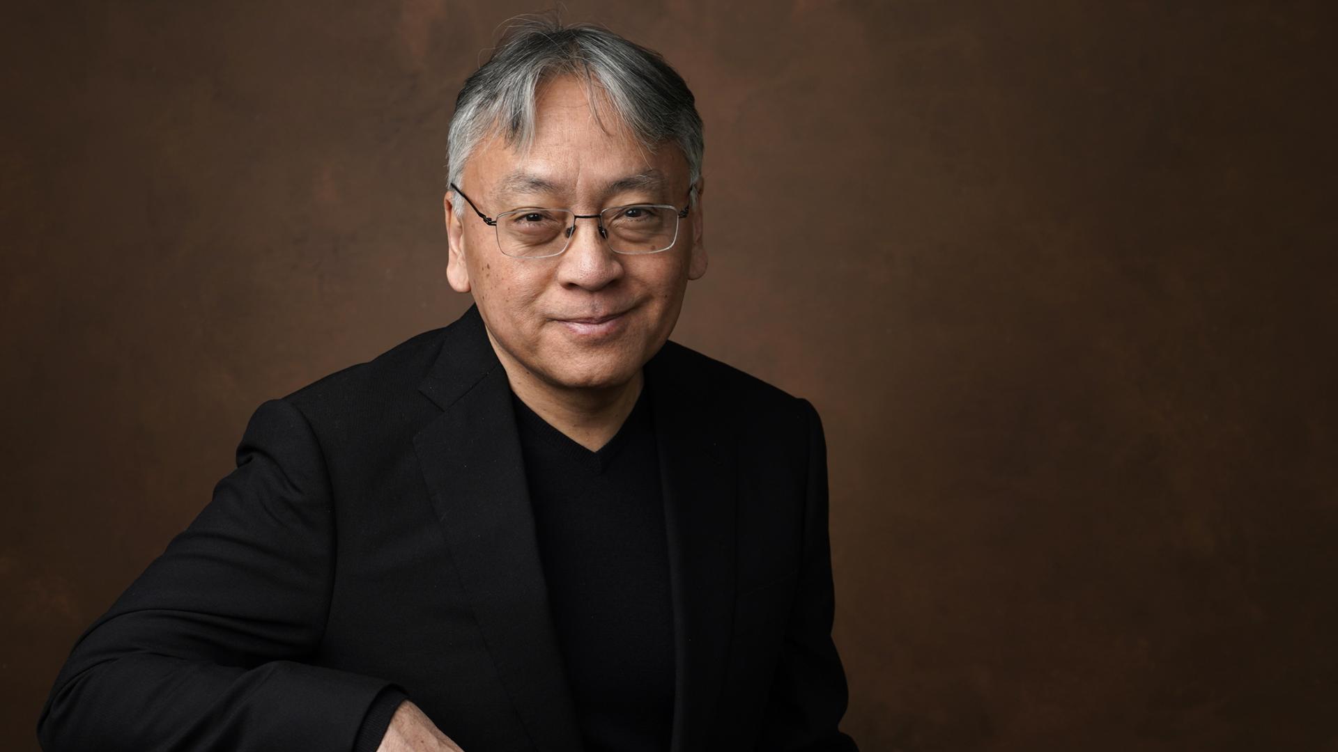 Kazuo Ishiguro sitzt vor neutralem Hintergrund und lächelt in die Kamera.