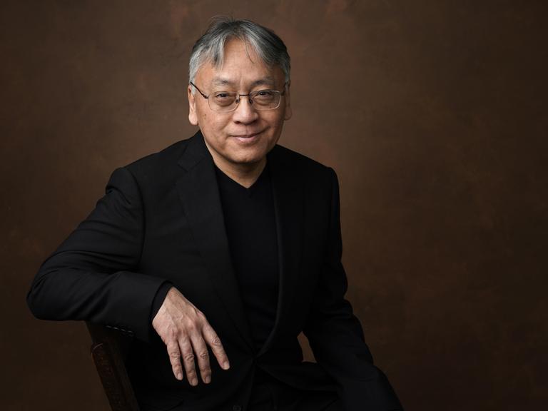 Kazuo Ishiguro sitzt vor neutralem Hintergrund und lächelt in die Kamera.