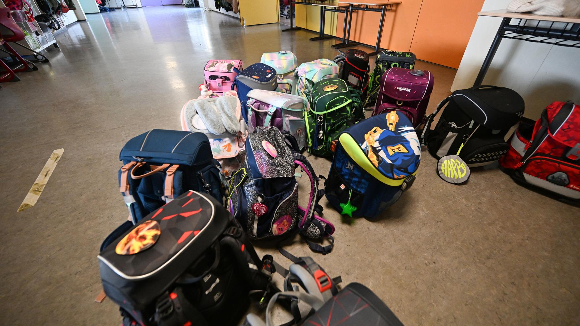 Bunte Schulranzen stehen und liegen in einer Grundschule vor einem Klassenzimmer auf dem Flur.