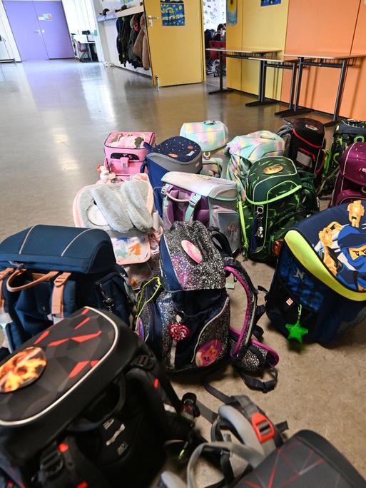 Bunte Schulranzen stehen und liegen in einer Grundschule vor einem Klassenzimmer auf dem Flur.