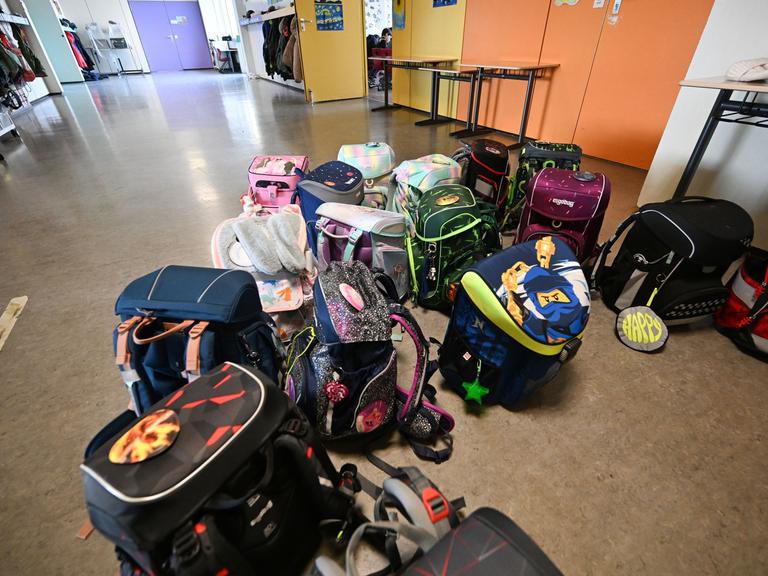 Bunte Schulranzen stehen und liegen in einer Grundschule vor einem Klassenzimmer auf dem Flur.