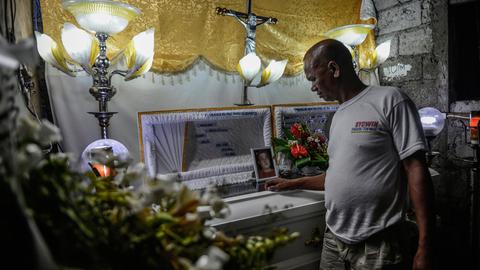 Ein Mann steht an einem Sarg, auf dem das Porträt eines jungen Mannes zu sehen ist. Ein Mann steht an einem Sarg, auf dem das Porträt eines jungen Mannes zu sehen ist.