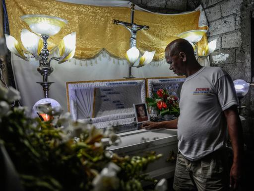 Ein Mann steht an einem Sarg, auf dem das Porträt eines jungen Mannes zu sehen ist.