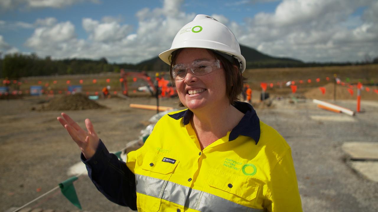 Eine junge Frau mit weißem Helm, Schutzbrille und gelber Warnjacke steht gestikulierend auf einer ebenerdigen Baustelle.