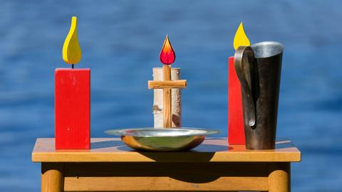 Beim Pfingstgottesdienst der Bremischen Evangelischen Kirche steht auf einem Tisch auf einem Platz vor der Weser ein Kreuz, ein Teller, ein Krug und Kerzenfiguren aus Holz. 
