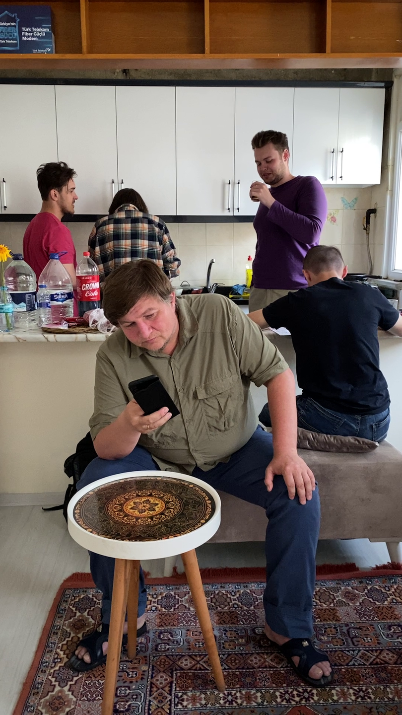 Ein Mann mittleren Alters sitzt auf einem Hocker und schaut in sein Handy. Hinter ihm werkeln jüngere Menschen in einer offenen Küche.