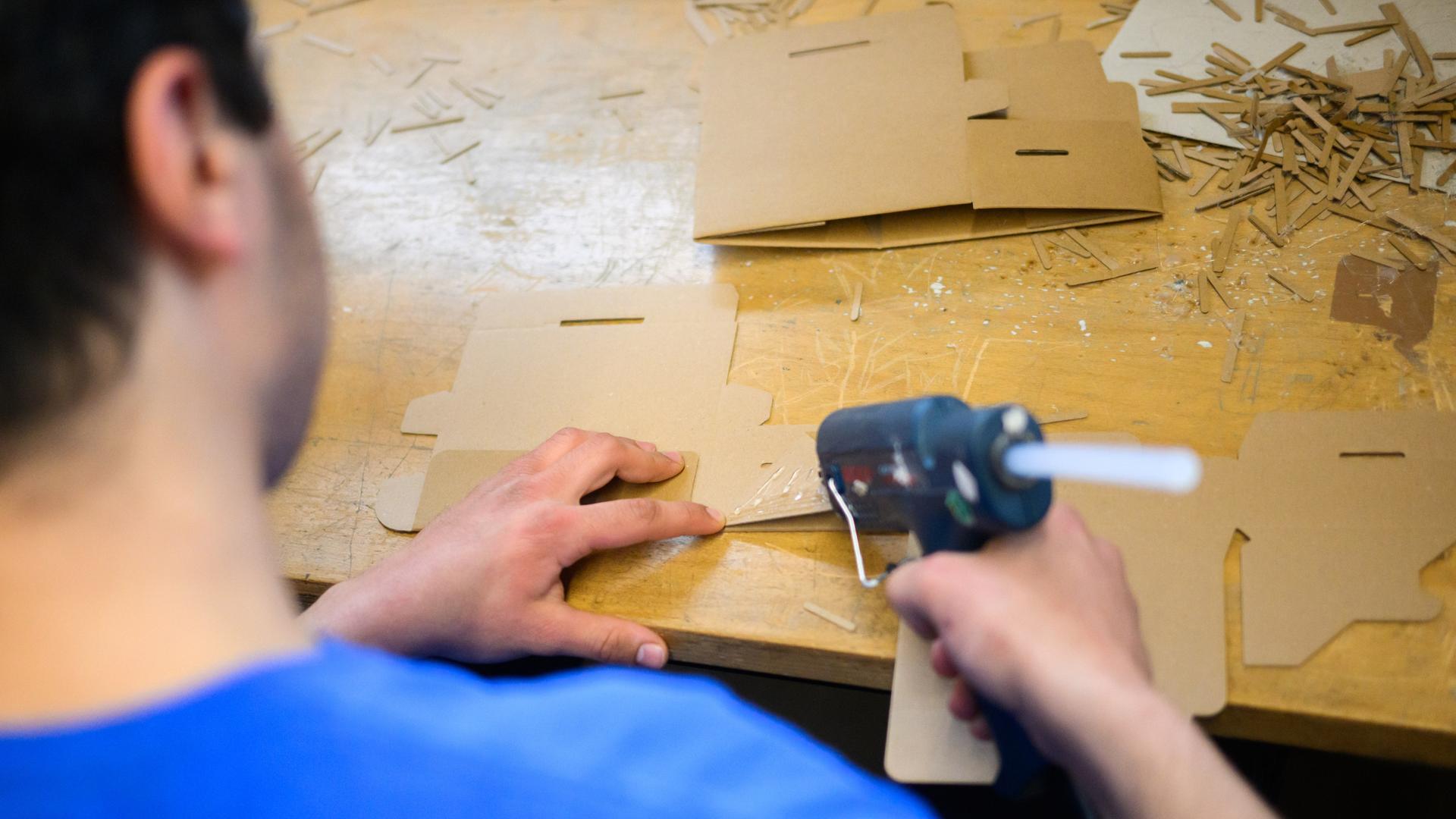 Ein junger Häftling klebt Kartons mit einer Klebepistole. Man sieht den Mann mit dunklen Haaren von hinten. Ein junger Häftling klebt Kartons mit einer Klebepistole. Man sieht den Mann mit dunklen Haaren von hinten.