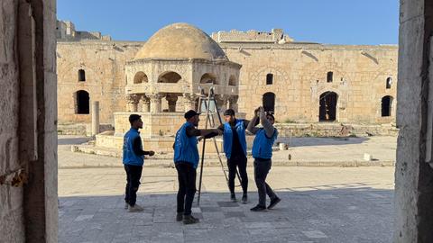 Syrische Arbeiter stehen im Hof einer Moschee und stellen eine Leiter auf. Sie tragen blaue Arbeitswesten.