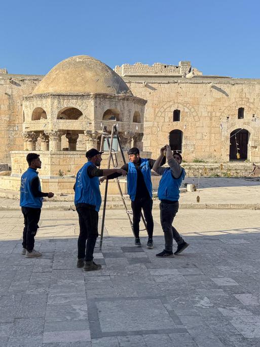 Syrische Arbeiter stehen im Hof einer Moschee und stellen eine Leiter auf. Sie tragen blaue Arbeitswesten.