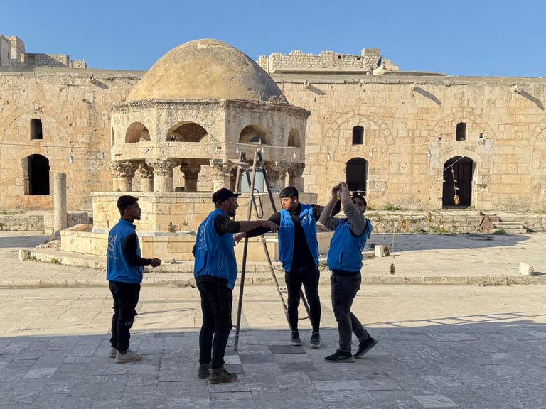 Syrische Arbeiter stehen im Hof einer Moschee und stellen eine Leiter auf. Sie tragen blaue Arbeitswesten.