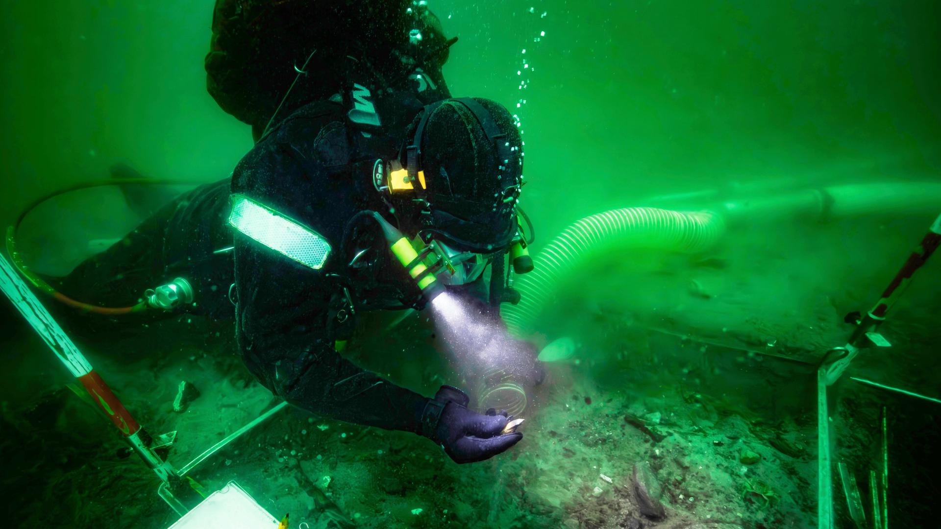 Das Bild zeigt einen Taucher, der unter Wasser archäologische Funde mit einer Taschenlampe untersucht. Das Bild zeigt einen Taucher, der unter Wasser archäologische Funde mit einer Taschenlampe untersucht.