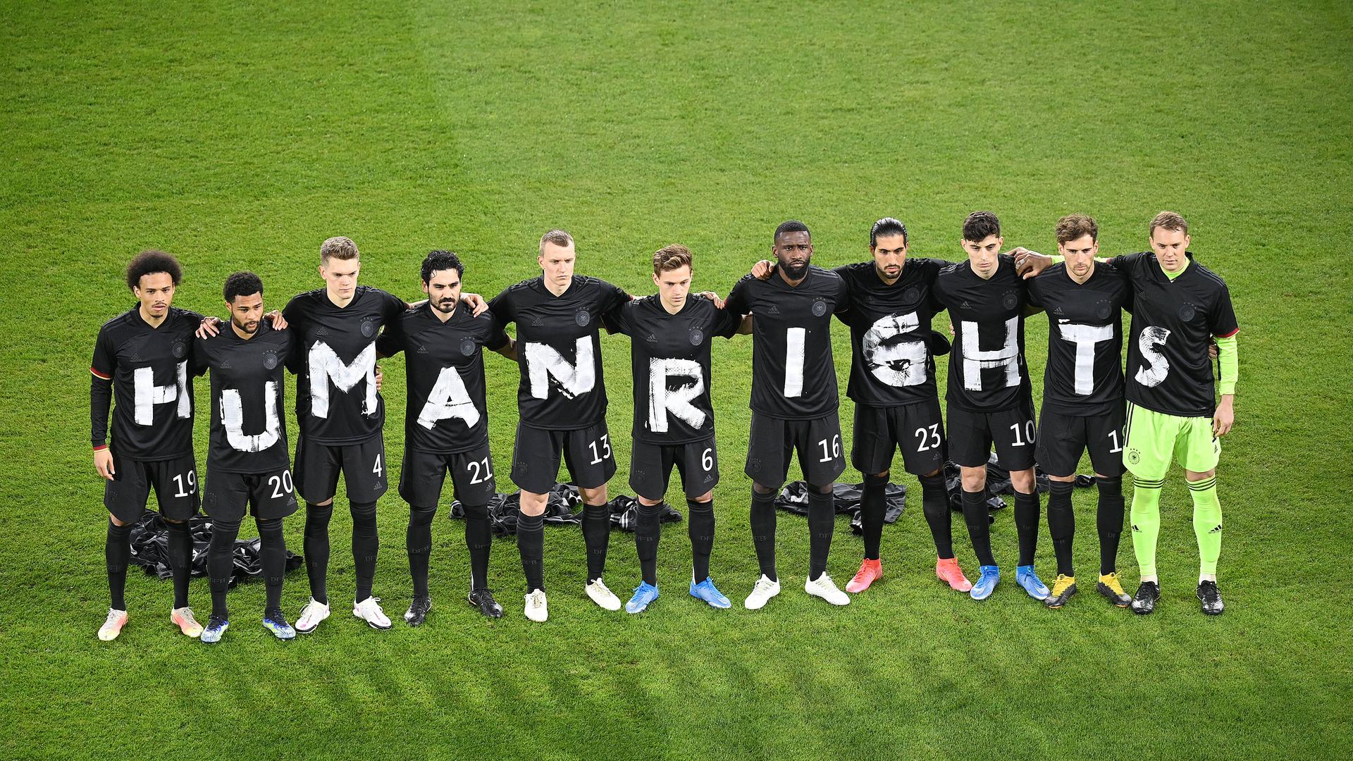 Fußballer stehen auf dem Rasen in einer Reihe und haben jeweils ein schwarzes T-Shirt an, auf dem mit weißer Farbe Human Rights geschrieben steht.
