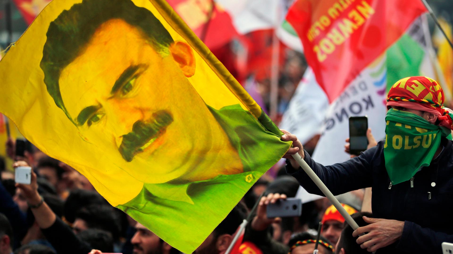 Ein Mann, der mit einem Tuch in den Farben der kurdischen PKK vermummt ist, hält eine Flagge mit dem Abbild von PKK-Gründer Öcalan in den Händen Ein Mann, der mit einem Tuch in den Farben der kurdischen PKK vermummt ist, hält eine Flagge mit dem Abbild von PKK-Gründer Öcalan in den Händen