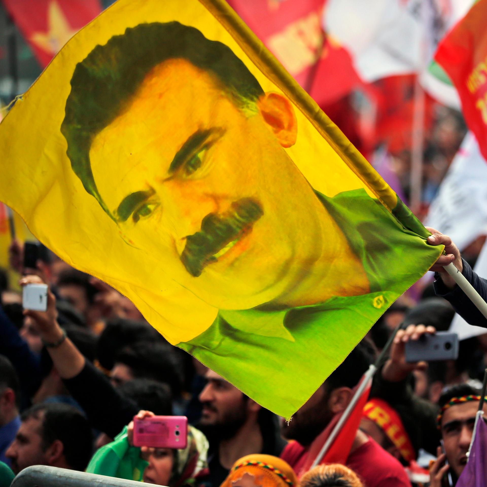 Ein Mann, der mit einem Tuch in den Farben der kurdischen PKK vermummt ist, hält eine Flagge mit dem Abbild von PKK-Gründer Öcalan in den Händen