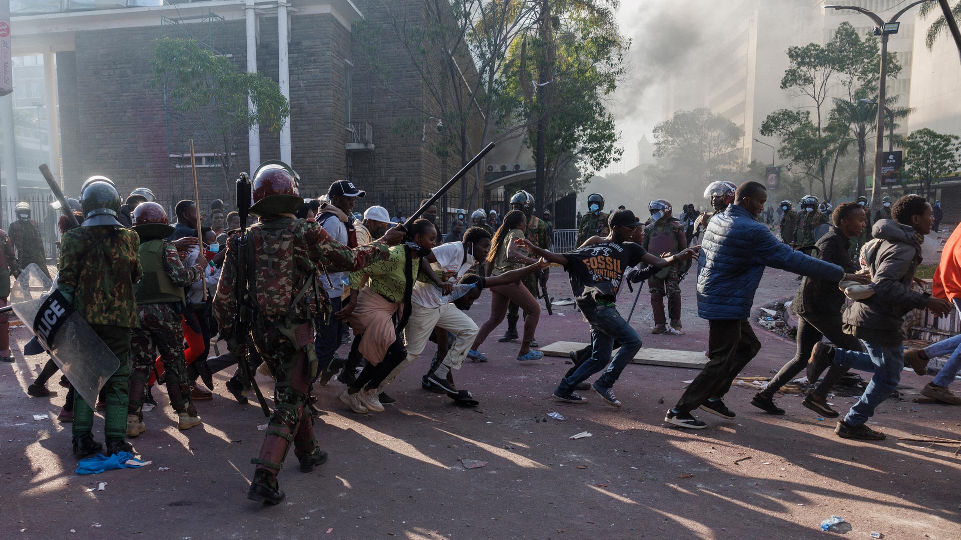Demonstrierende Männer und Frauen halten sich in Nairobi an den Händen und laufen an zwei schwer bewaffneten Polizisten vorbei. Im Hintergrund steigt Rauch auf.