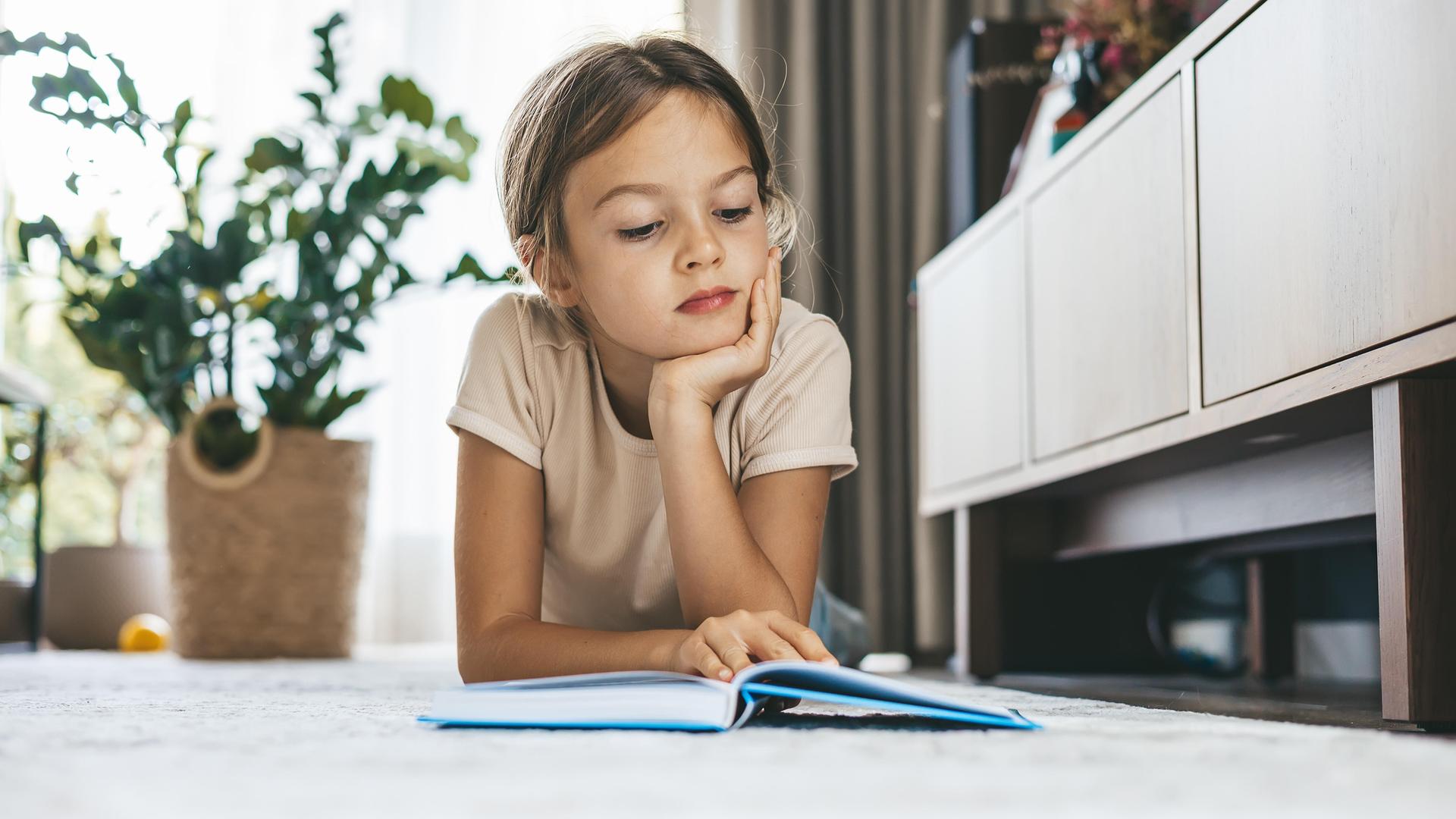 Ein junges Mädchen liegt im Wohnzimmer auf dem Boden und liest ein Buch.