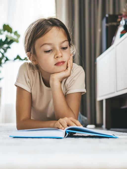 Ein junges Mädchen liegt im Wohnzimmer auf dem Boden und liest ein Buch. Ein junges Mädchen liegt im Wohnzimmer auf dem Boden und liest ein Buch.