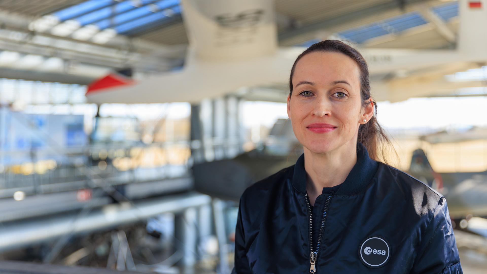 Nicola Winter trägt eine Jacke mit der Aufschrift "esa" und steht in einer Halle, in der im Hintergrund Flugzeuge zu sehen sind