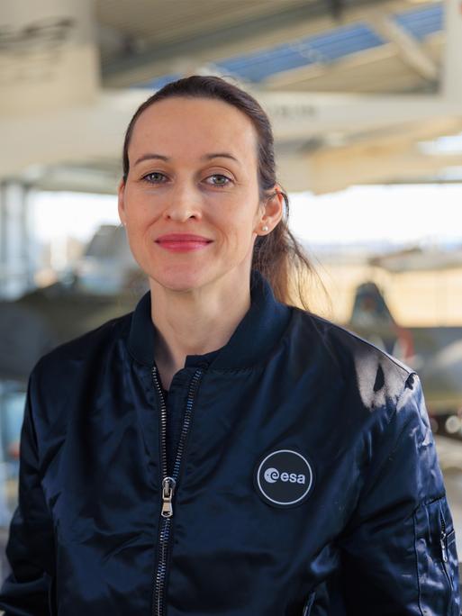 Nicola Winter trägt eine Jacke mit der Aufschrift "esa" und steht in einer Halle, in der im Hintergrund Flugzeuge zu sehen sind