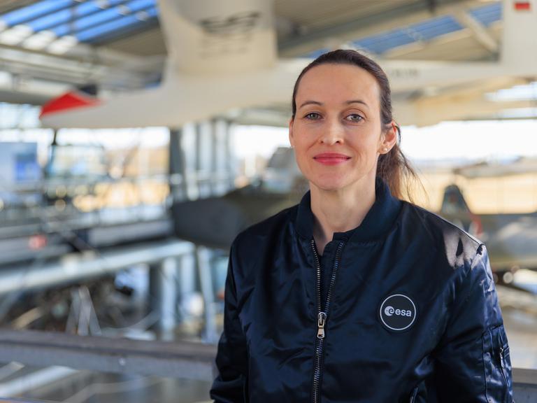 Nicola Winter trägt eine Jacke mit der Aufschrift "esa" und steht in einer Halle, in der im Hintergrund Flugzeuge zu sehen sind