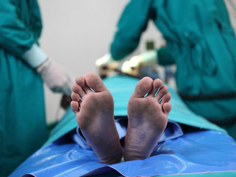 Füße in Nahaufnahme auf einem Behandlungstisch in einem Krankenhaus. Im Hintergrund sieht man zwei Menschen in OP-Kleidung bei vermutlich lebenserhaltenden Maßnahmen.