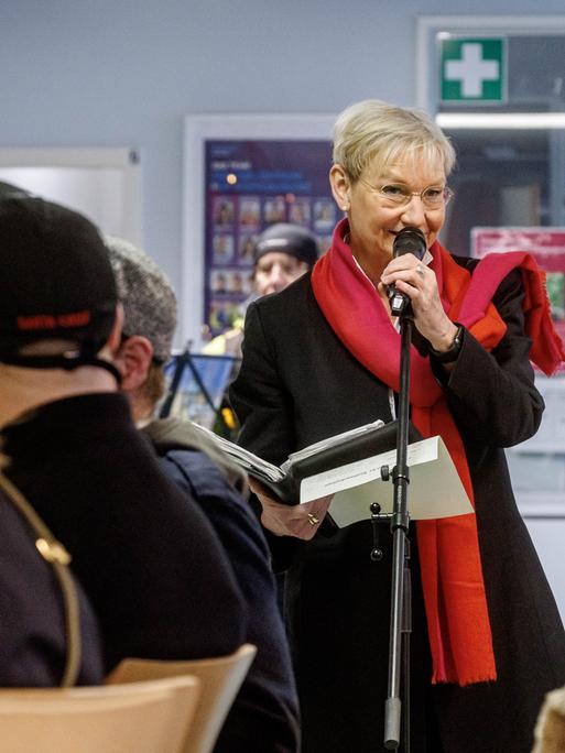 Kirsten Fehrs steht in einem Raum der Diakonie in Hamburg. Vor ihr sitzen Menschen an Tischen. Sie trägt einen schwarzen Mantel und einen roten Schal und spricht in ein Mikrofon.