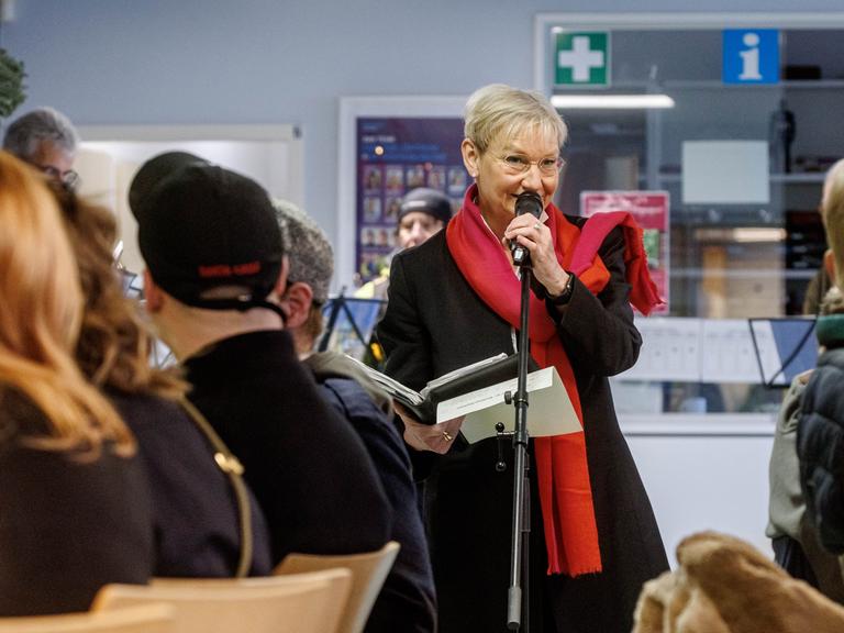 Kirsten Fehrs steht in einem Raum der Diakonie in Hamburg. Vor ihr sitzen Menschen an Tischen. Sie trägt einen schwarzen Mantel und einen roten Schal und spricht in ein Mikrofon.