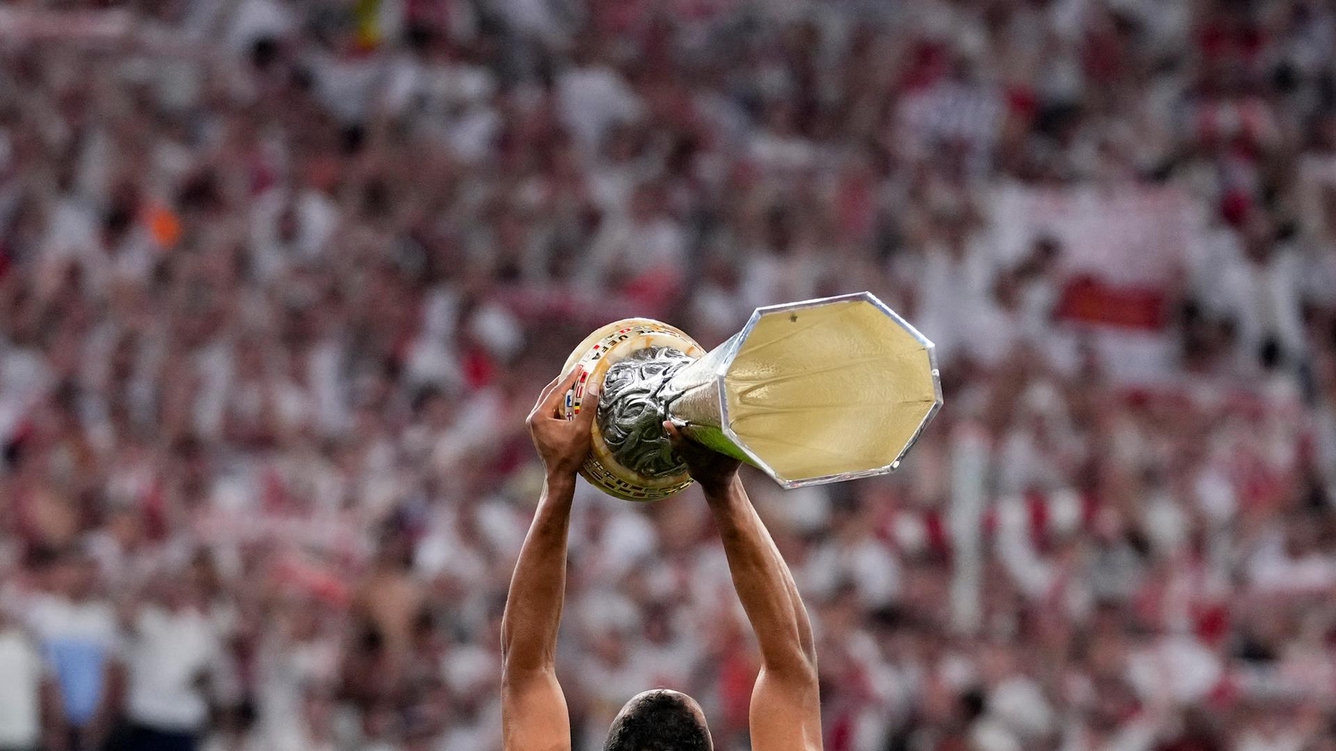 Ein Mann in weißem Fußball-Trikot ist von hinten zu sehen. Er hält den silbernen Pokal mit beiden Händen in die Höhe und präsentiert ihn einem großen Publikum, das aber nur schemenhaft im Hintergrund auf den Rängen und Tribünen des Stadions in der ungarischen Hauptstadt Budapest zu sehen ist.