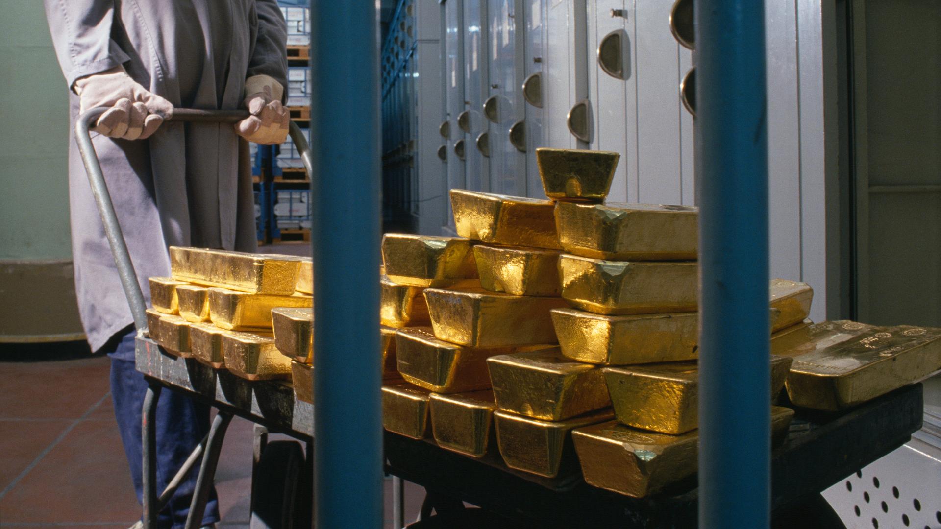 Gestapelte Goldbarren der "Banque de France" werden im Jahr 1989 von einem Bankmitarbeiter mit Handschuhen und grauem Arbeitskittel auf einem Karren hinter Gittern geschoben. Im Hintergrund stehen viele Metallschränke und Regale. Gestapelte Goldbarren der "Banque de France" werden im Jahr 1989 von einem Bankmitarbeiter mit Handschuhen und grauem Arbeitskittel auf einem Karren hinter Gittern geschoben. Im Hintergrund stehen viele Metallschränke und Regale.