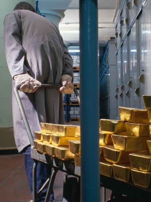 Gestapelte Goldbarren der "Banque de France" werden im Jahr 1989 von einem Bankmitarbeiter mit Handschuhen und grauem Arbeitskittel auf einem Karren hinter Gittern geschoben. Im Hintergrund stehen viele Metallschränke und Regale.