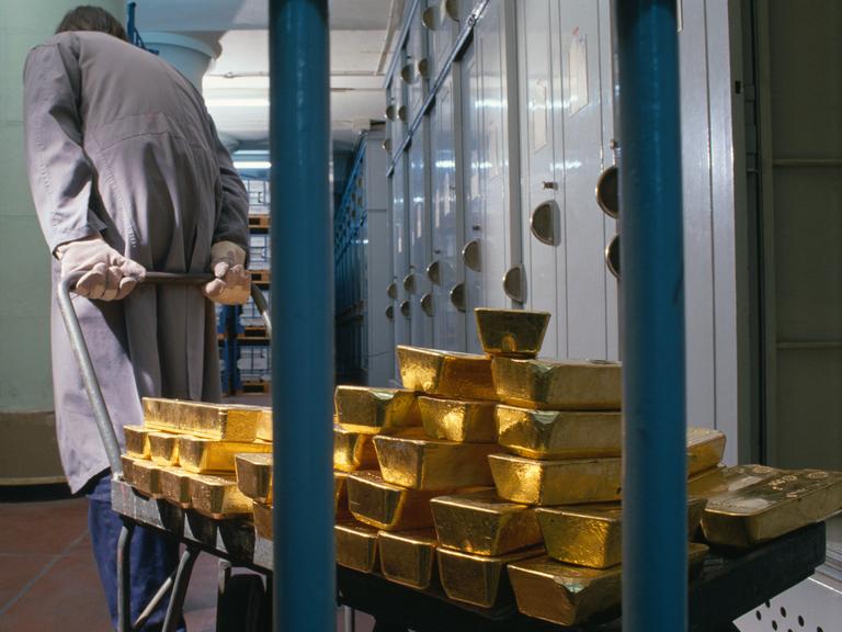 Gestapelte Goldbarren der "Banque de France" werden im Jahr 1989 von einem Bankmitarbeiter mit Handschuhen und grauem Arbeitskittel auf einem Karren hinter Gittern geschoben. Im Hintergrund stehen viele Metallschränke und Regale.