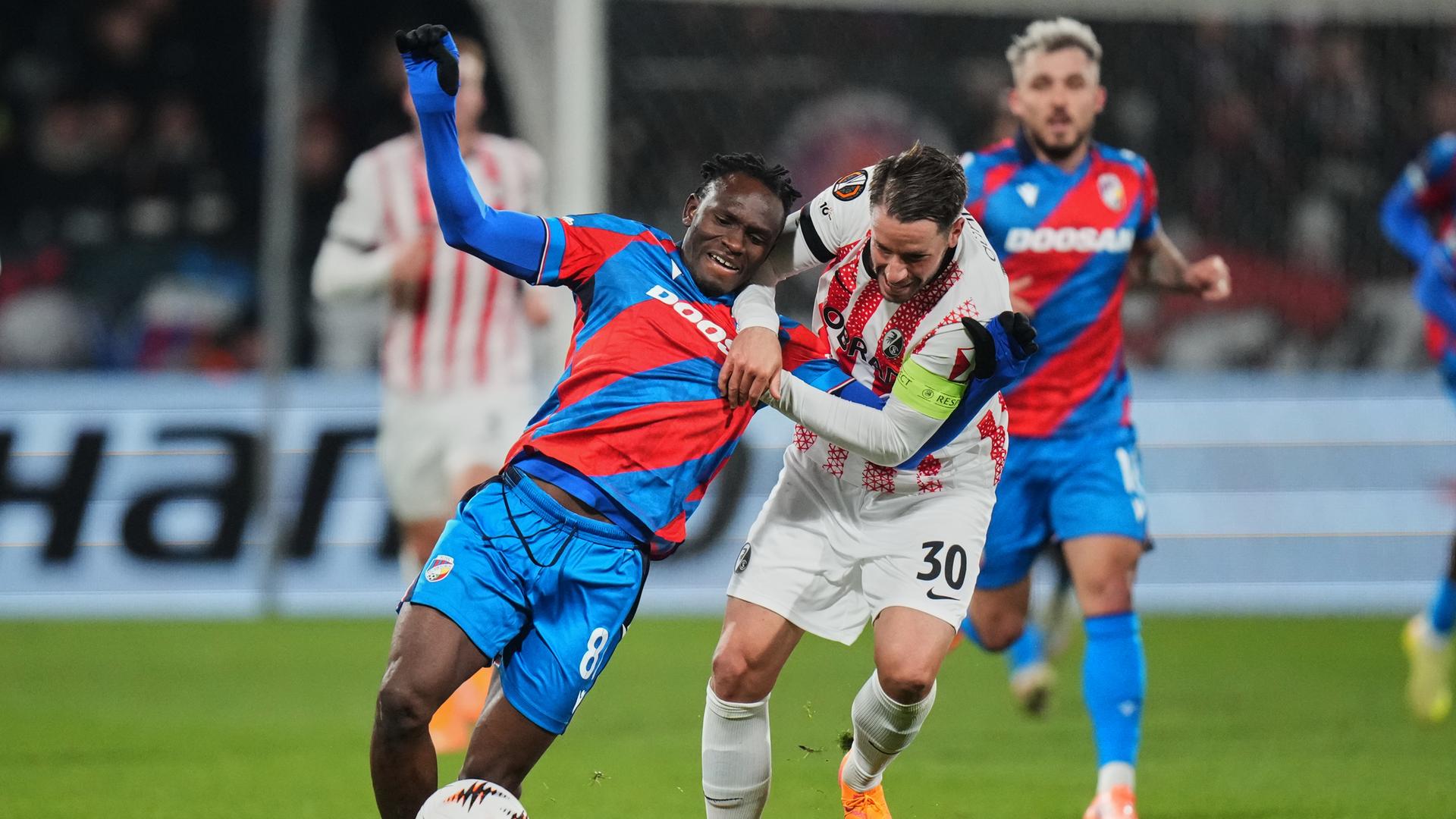 Die beiden laufen nebeneinander her. Günther zieht an Adus Oberkörper, der umzufallen scheint. Günther trägt ein weißes Trikot, Adu ein schräg blau-rot gestreiftes.