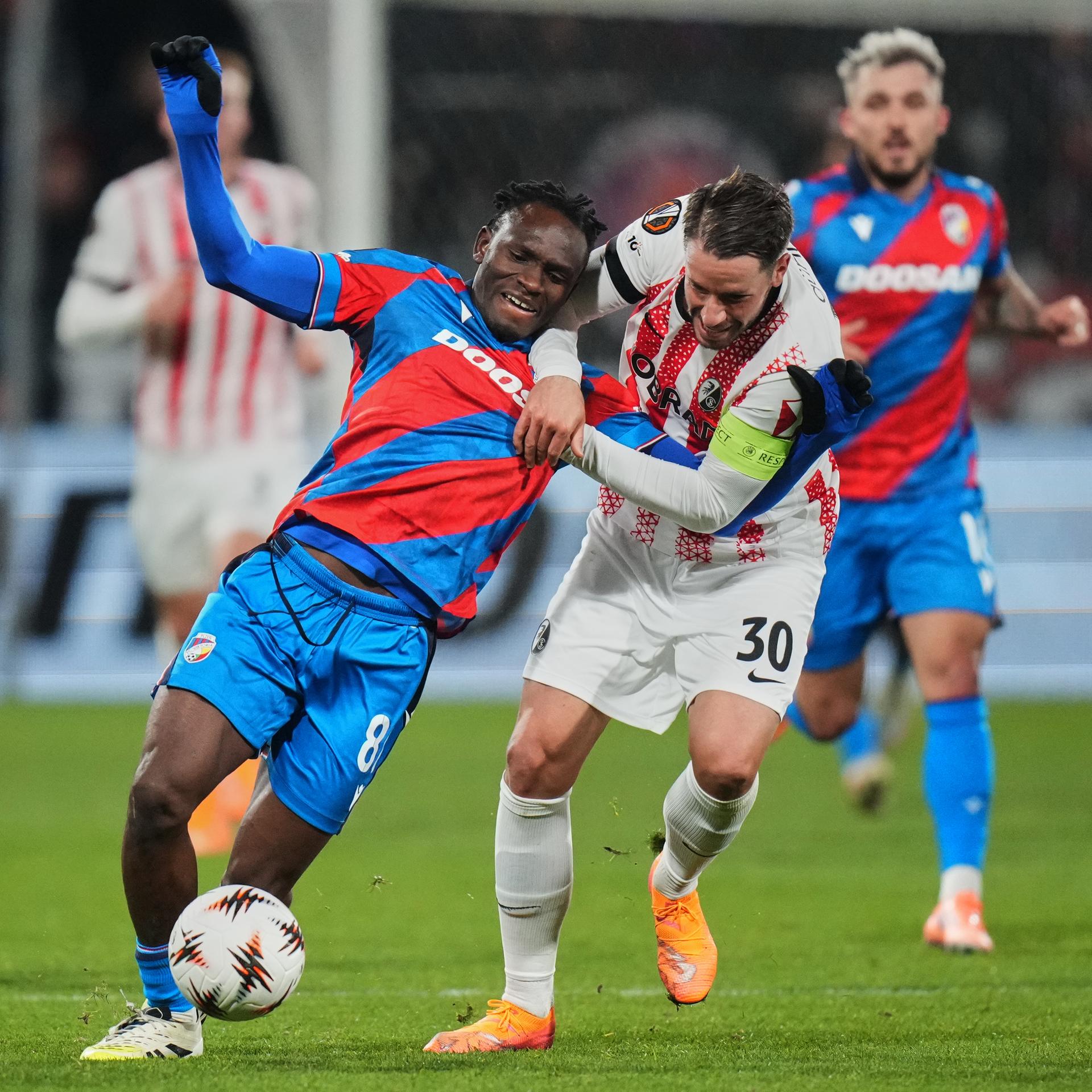 Die beiden laufen nebeneinander her. Günther zieht an Adus Oberkörper, der umzufallen scheint. Günther trägt ein weißes Trikot, Adu ein schräg blau-rot gestreiftes.