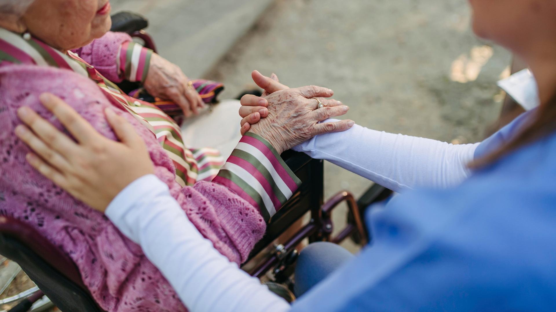 Eine Altenpflegerin hält die Hand einer alten Dame, die im Rollstuhl vor ihr sitzt.
